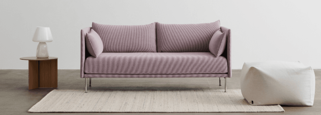 Modern pink Silhouette Sofa with pillows and metal legs, on a rug with a side table and ottoman.