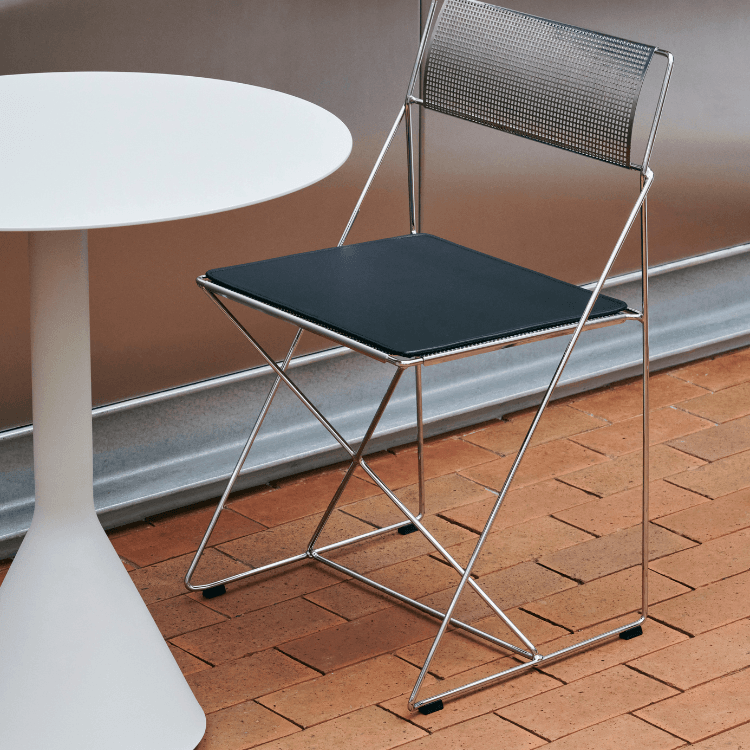 Modern X-Line Chair Chrome with black seat and chrome frame, next to a white table, on a brick surface.