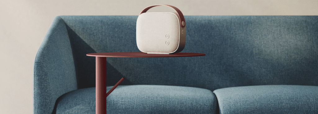 AUTO-DRAFT speaker in cream with a brown handle, sitting on a red side table next to a blue sofa.