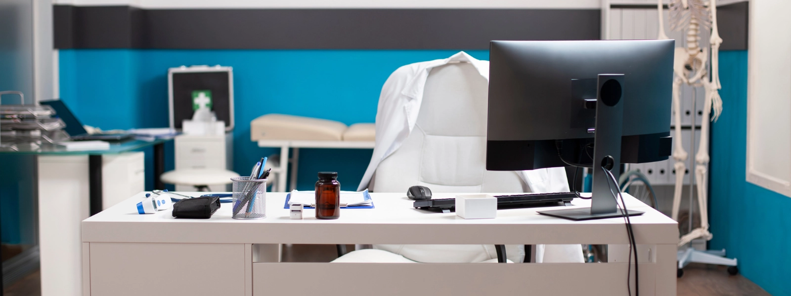 Healthcare workstation with doctor's coat on chair, computer, medical supplies, and skeleton model.