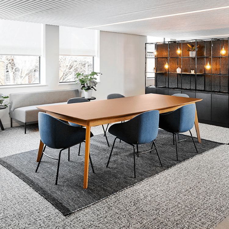 Dalby Table with a warm wood finish and slender profile, surrounded by blue upholstered chairs in a modern office space.