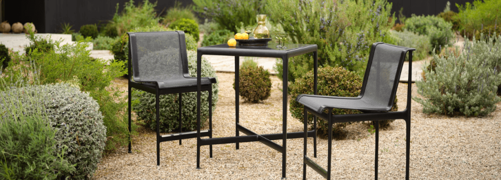 Pair of black 1966 Stool chairs and a matching table placed on a garden gravel path with lush green shrubbery.