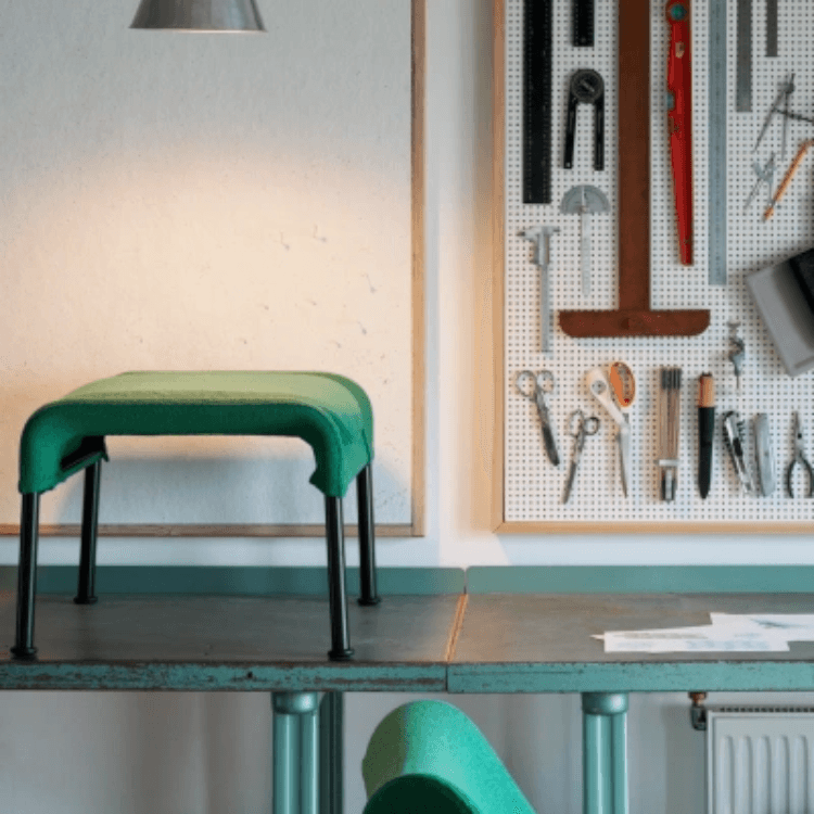 Green O2 Ottoman with black legs placed on a workspace table next to a pegboard of tools.