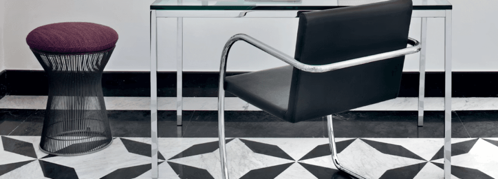 Platner Stool with dark steel wire base and textured berry-colored cushion placed beside a sleek glass-topped desk.