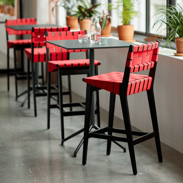 Risom Barstool with a black hardwood frame and vibrant red woven seat, positioned at a sleek table in a bright room.