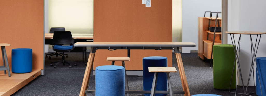 Rockwell Unscripted Easy Stools paired with colorful fabric cylinders around a central wood and metal workstation table.